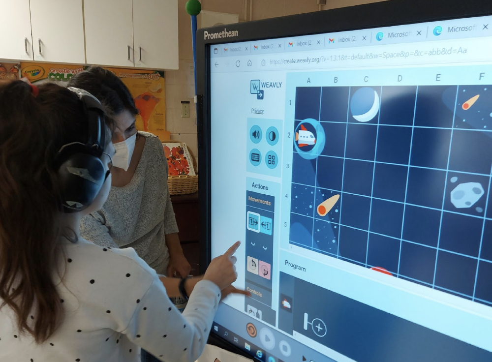 A student and a facilitator building a program in the Weavly space theme using a Promethean board.