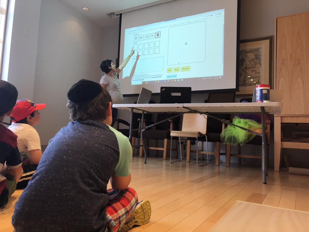 A facilitator explains the aspects of the prototype while it is projected on the screen to campers who are seated on the ground watching and listening.