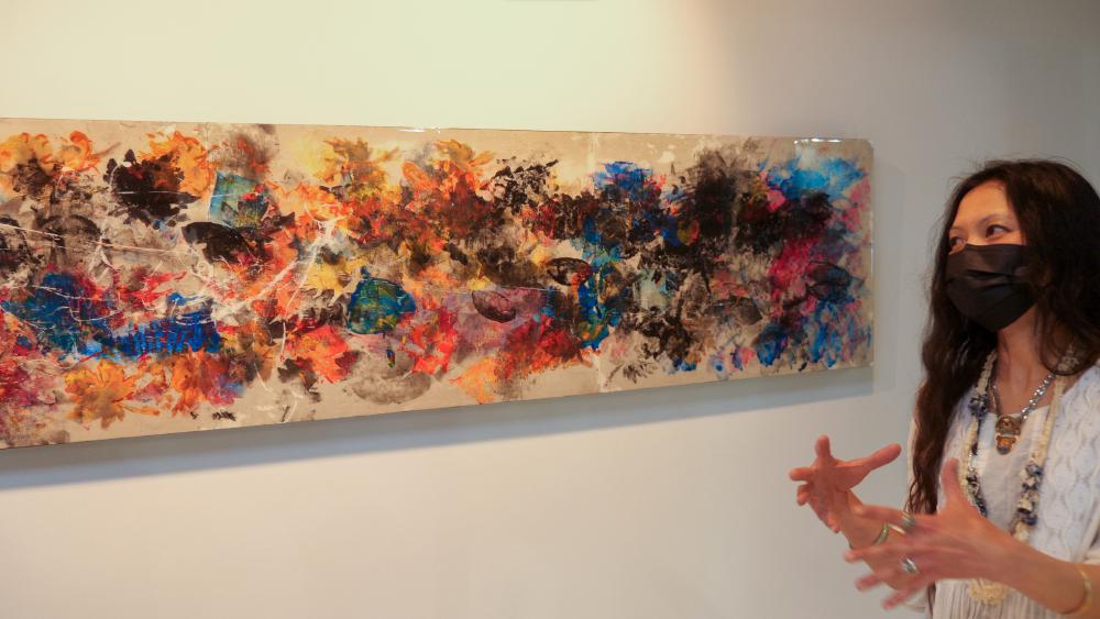 An abstract, colourful painting with impressions of leaves on it. A woman is standing to the right describing the painting.