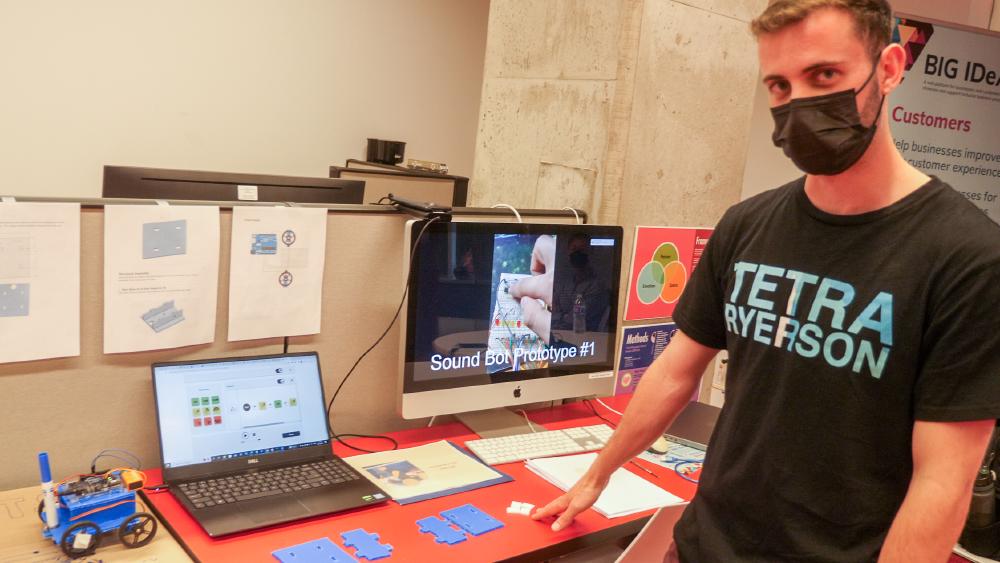 A man stands in front of a computer showing a prototype of a Sound Robot. Beside the computer is a simple DIY robot.