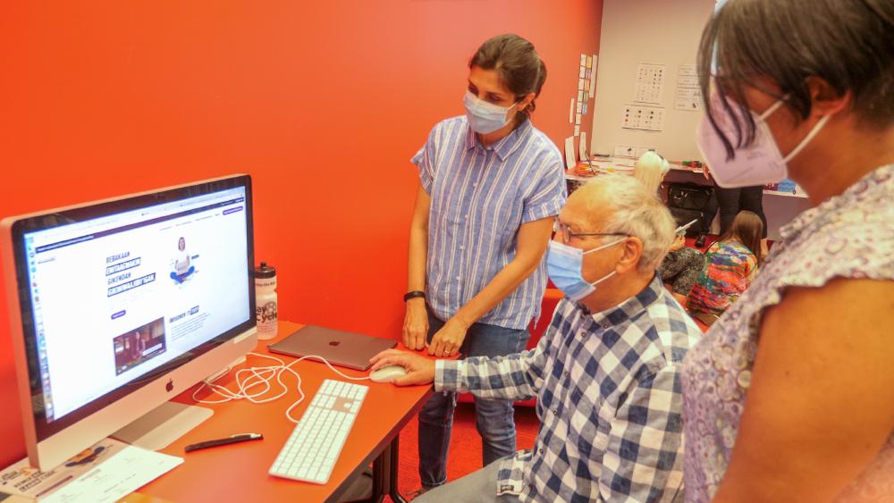 Three people are looking at the Your Voice is Power website. In the background, children are playing with robots on the floor.