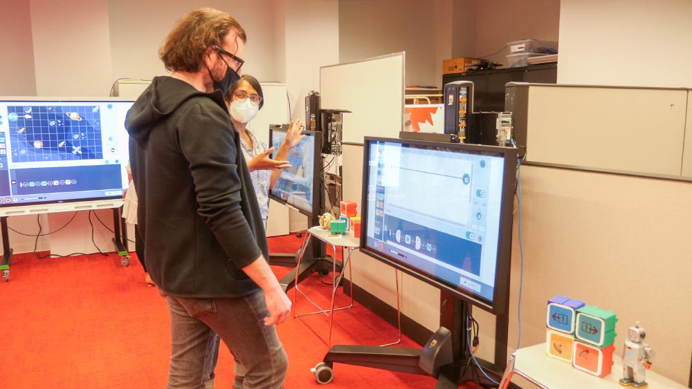 Two people stand in front of a smart board which has the Weavly coding environment on it. In the foreground there are large colourful blocks with the Weavly actions on them and a toy robot.